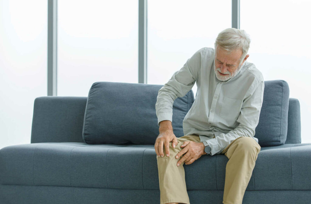 An elderly man that is sitting on the sofa is experiencing joint pain and having problems standing by himself.