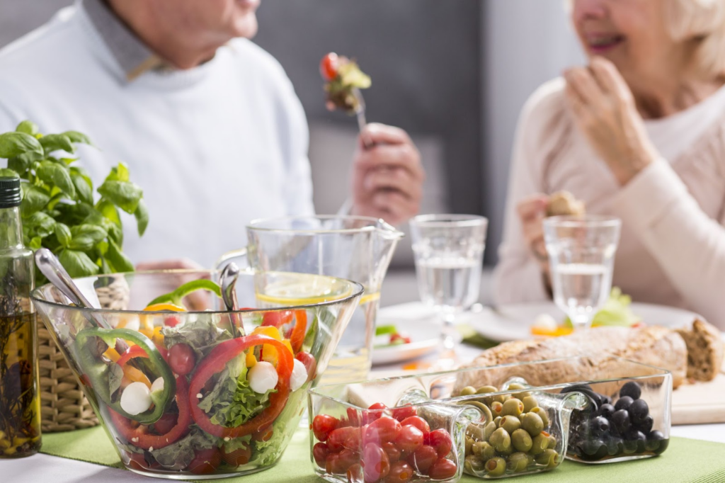 An elderly couple in the background eating a salad with olives, tomatoes and bread on the table.