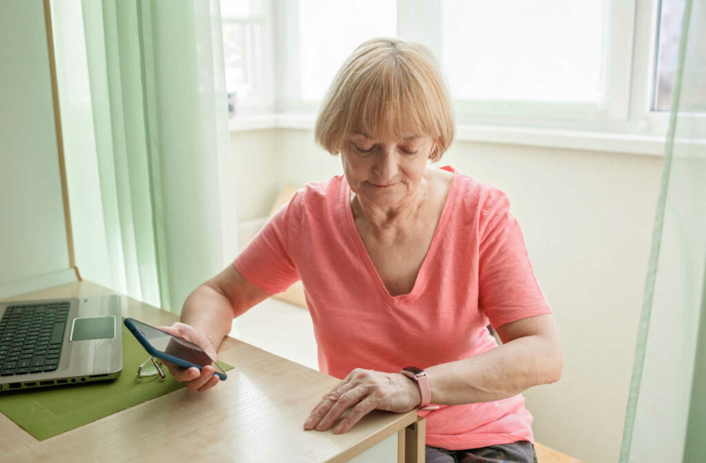 A senior woman with a laptop on a desk, watching her smartwatch while it’s being connected to her smartphone.