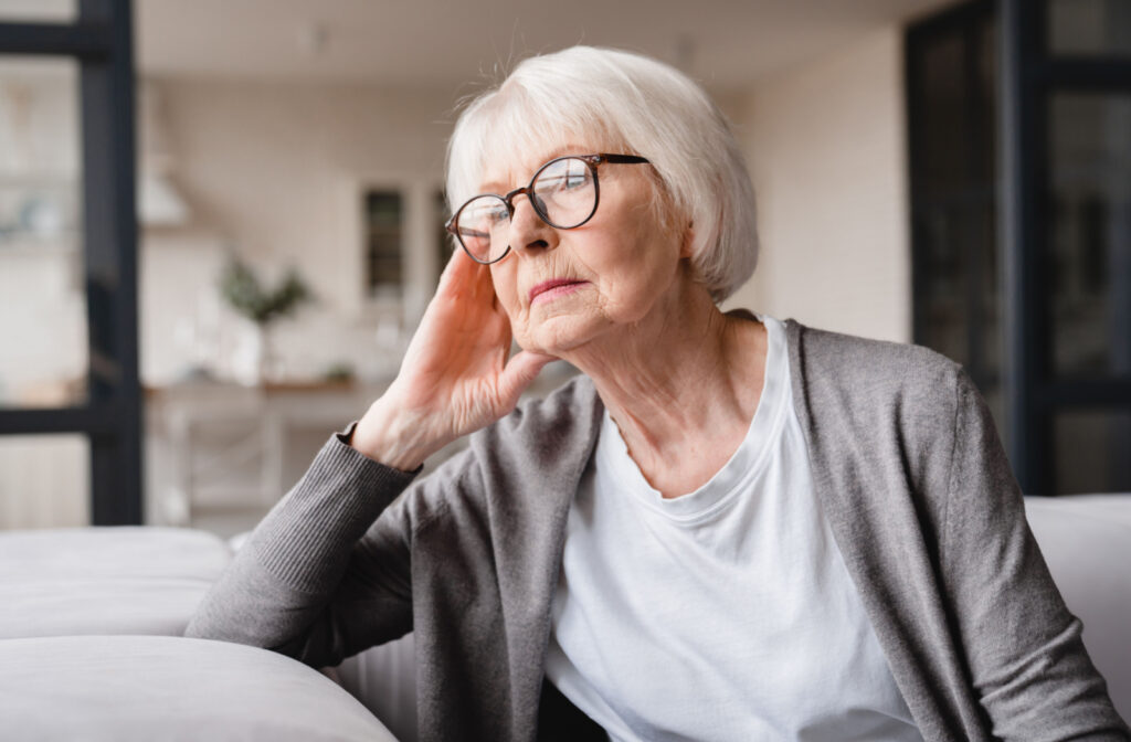 A senior woman with glasses on sitting on a couch and looking out the window with a serious expression