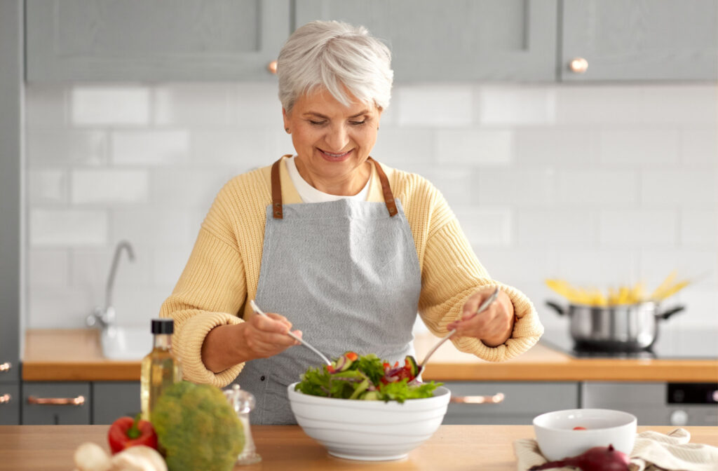 A senior woman in the kitchen preparing a bowl of salad using both hands and smiling