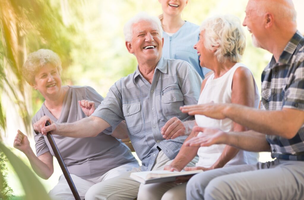 An elderly man with a cane smiles and laughs while sitting outside with a nurse and other older adults.