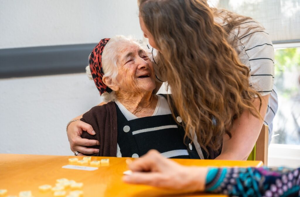 An adult child hugs their older parent with dementia in the middle of playing a memory game together with a friend