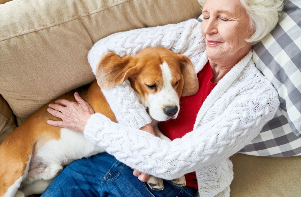 A senior wearing a white sweater smiles gently while they cuddle their pet beagle on a beige-colored couch