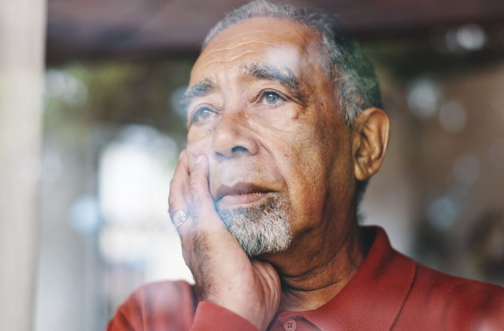 Elderly person sitting by a window with a hand on their face appearing deep in thought