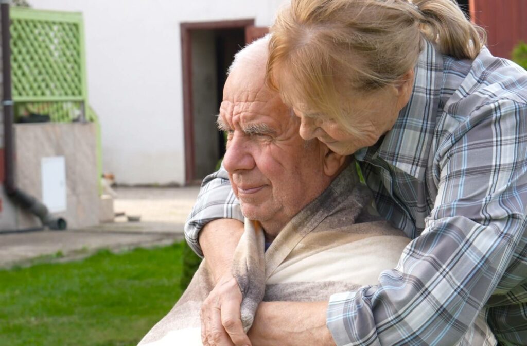 Elderly person wrapped in a blanket in a wheelchair receiving support from another individual in an outdoor setting