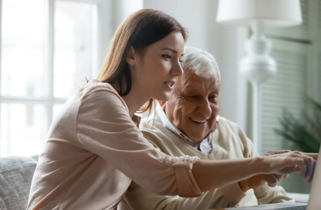 An adult child helps their older parent research senior living communities, pointing at the screen and laughing