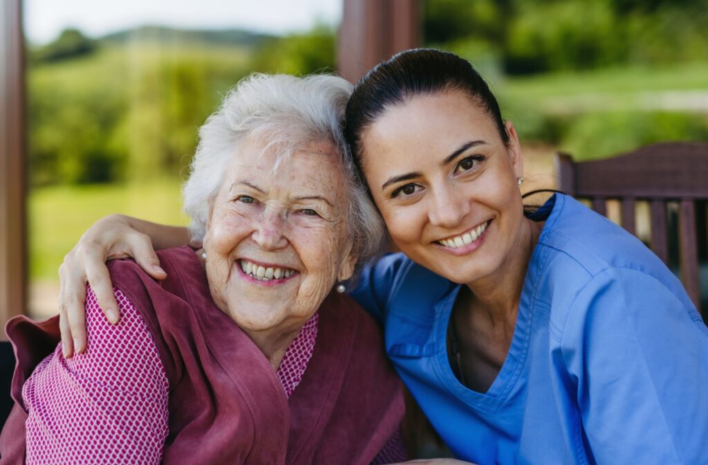 A caregiver and an older adult grin at the camera while sitting on a bench together in senior living