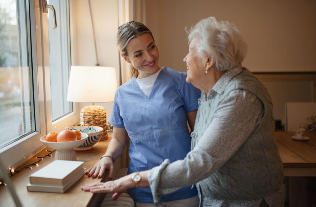 A caregiver smiles at an older adult while they stand beside a desk in front of a window in their home in memory care